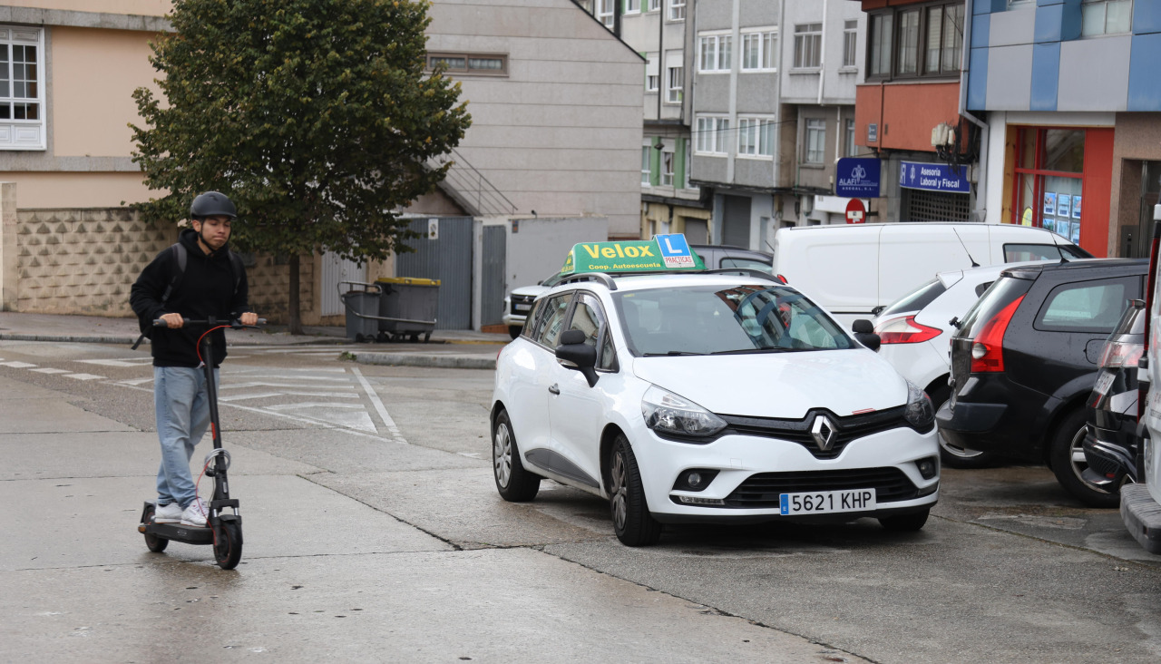 Un conductor de patinete circulando al lado de un coche de autoescuela