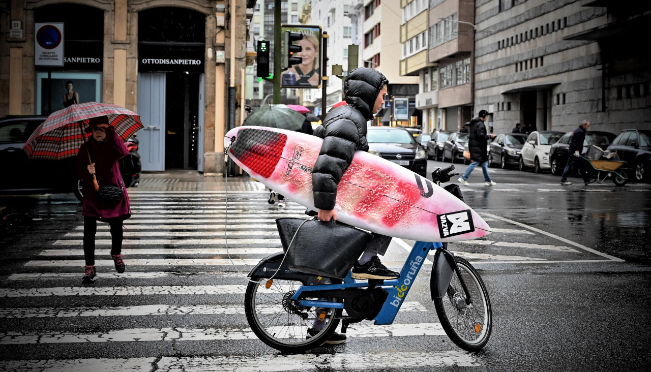 Un usuario de BiciCoruña, con una tabla de surf en la mano en medio de una jornada lluviosa